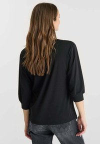 Donna con lunghi capelli castani che indossa un top nero a tre quarti e jeans grigio scuro, voltata di spalle su uno sfondo neutro.