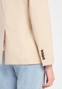 Beige Blazer mit einem Schlitz hinten und fünf dunklen Knöpfen an den Ärmeln, kombiniert mit hellblauen Jeans.