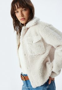 Faux shearling jakke i lys beige med krave, knaplukning, to brystlommer og en tekstureret overflade. Båret over en cremet top.