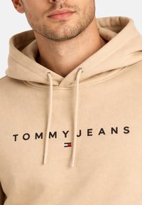 Uomo che indossa una felpa con cappuccio beige con cordini e scritta "TOMMY JEANS" con un piccolo logo rosso, bianco e nero sul petto.