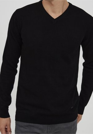 Man draagt een effen zwarte V-hals trui met lange mouwen en grijze broek, staand tegen een effen achtergrond.