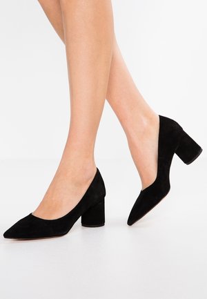 Une paire de jambes portant des chaussures en daim noir à talon bas bloc et bout pointu, sur fond blanc.