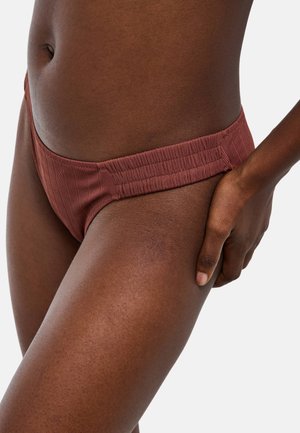 Gros plan d'une personne portant un bas de bikini marron côtelé avec des côtés élastiques larges, main reposant sur la cuisse supérieure sur un fond clair.