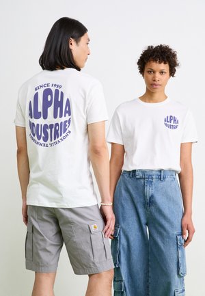 Hvite bomulls t-skjorter med lilla "ALPHA INDUSTRIES" trykk, en med stort design på baksiden og en med lite logo på brystet, kombinert med uformelle shorts og jeans.
