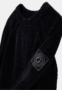 Schwarzer, strukturierter Pullover mit geripptem Rundhalsausschnitt und einem Stoffpatch am Ärmel, das ein kreisförmiges Logo und Textdetails zeigt.