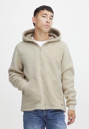 Beige fleece hoodie met een volledige ritssluiting, capuchon met trekkoord en twee voorzakken. Zachte textuur en geribbelde manchetten. Casual ontwerp in een relaxte pasvorm.