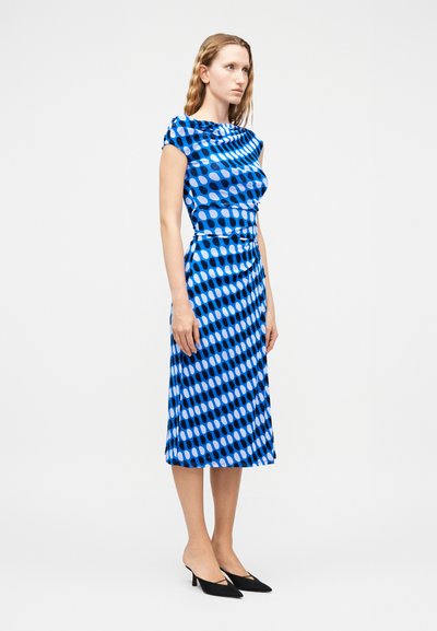 Γυναίκα που στέκεται φορώντας ένα μπλε μίντι φόρεμα με σχέδιο από μαύρα και ανοιχτά μπλε στρογγυλεμένα ορθογώνια και μαύρα μυτερά παπούτσια με χαμηλό τακούνι.