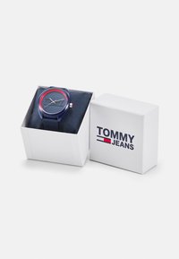 Reloj azul con una esfera redonda, acentos rojos y un logo de Tommy Jeans, presentado en una caja blanca con forro de tela azul.