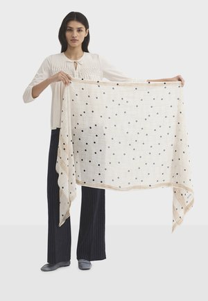 Frau trägt eine weiße Bluse und schwarze Hosen und hält einen großen beigen Schal mit schwarzen Punkten und einem feinen Karomuster.