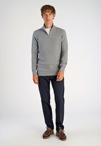 Hellgrauer Pullover mit Reißverschluss und langen Ärmeln, kombiniert mit blauen Hosen und braunen Schnürschuhen. Einfacher, strukturiertes Gewebe.