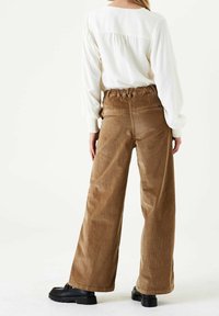 Brown corduroy wide-leg pants with an elastic waist, featuring two back pockets. Worn with a long-sleeve white top and black shoes.