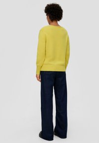 Geel gebreid trui met een ronde hals en geribde zoom, gecombineerd met donkerblauwe wijde jeans en zwarte schoenen, gezien vanaf de achterkant.