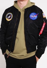 Giacca bomber nera con toppe NASA e missioni spaziali, chiusura con zip, polsini a costine e un accento con portachiavi rosso. Indossata sopra un hooded verde.
