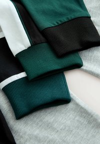 Drei gefaltete Langarmshirts in Grün, Schwarz, Weiß und Grau mit gerippten Bündchen, auf einer hellen Oberfläche gestapelt.