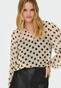 Gennemsigtig beige bluse med sorte prikker, har en V-hals og lange ærmer, parret med en sort lædernederdel.