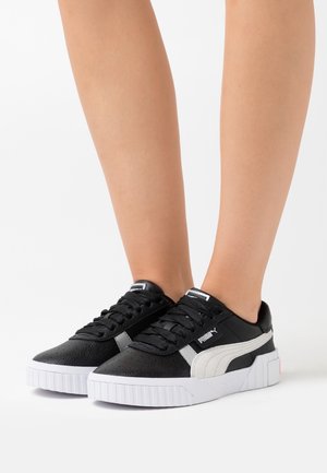 Jambes portant des baskets Puma noires avec des semelles blanches et des rayures latérales, debout sur une surface blanche.