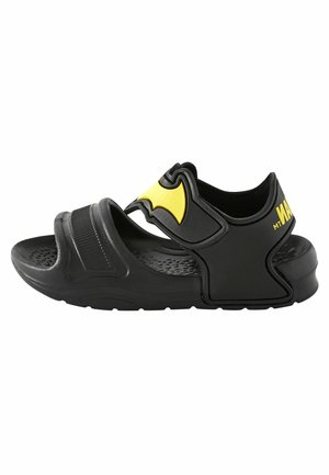 Schwarze Sandalen mit einer strukturierten Sohle, verstellbarem Riemen mit einem gelben Fledermaus-Emblem und einem gepolsterten, konturierten Design für Komfort.