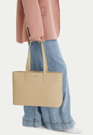 Osoba v širokých modrých džínách a růžovém kabátě drží béžovou tašku přes rameno s dvojitými uchy a malým logem.