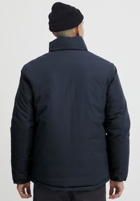 Mann, der eine dunkelblau gepolsterte Jacke und eine schwarze Strickmütze trägt, von hinten vor einem schlichten hellen Hintergrund gezeigt.