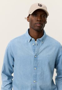 Ανοιχτό μπλε πουκάμισο από τζιν με γιακά που κουμπώνει και λογότυπο στο στήθος. Μπεζ καπέλο μπέιζμπολ με λογότυπο «LD». Απλός, καθημερινός σχεδιασμός.