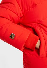 Czerwona puchowa kurtka z miękkim materiałem zewnętrznym, elastycznymi mankietami oraz małą naszywką z logo na rękawie. Gruby, pikowany materiał zapewniający izolację.