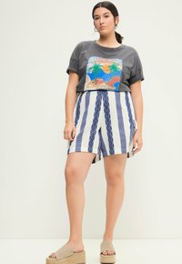 Šedé grafické tričko s tropickým designem, spárované s modro-bílými pruhovanými šortkami s geometrickými vzory. Nosené s béžovými platformovými sandály.
