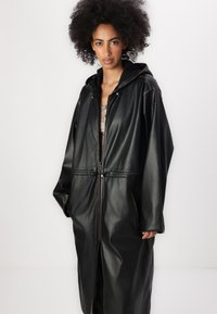 Caban oversized din piele ecologică neagră cu glugă, dotat cu un fermoar frontal, talie elastică și două buzunare laterale. Textură netedă, cusături minime.