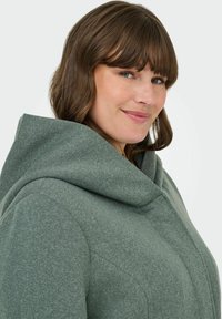 Manteau à capuche vert en tissu texturé, avec un large col et des détails de couture. Le design comprend une coupe décontractée.
