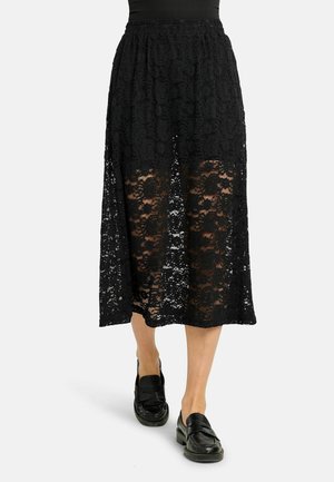 Čierna čipkovaná sukňa strednej dĺžky s kvetinovým vzorom, kombinovaná s čiernymi mokasínami a vsadeným čiernym topom, na svetlosivom pozadí.