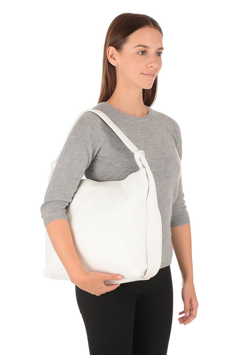Witte leren tote bag met een brede body, enkele schouderband en een lichte glans. Model houdt hem tegen een grijze trui en zwarte broek.