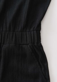 Zwart textuurstof kledingstuk met geplooide elastische tailleband en zichtbare zijnaadzak op een lichte achtergrond.