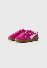 Un paio di sneakers Puma in suede rosa con strisce bianche sui lati, suole in gomma, chiusura con lacci e piccoli charm decorativi sui lacci.