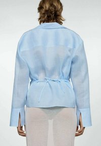 Camicia in lino azzurro chiaro, semi-trasparente, con vita regolabile, maniche lunghe e spacchi laterali, dotata di colletto classico e vestibilità rilassata.