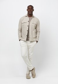 Giacca camicia in lino beige chiaro con due tasche sul petto, indossata sopra una maglietta grigia, abbinata a pantaloni bianchi e sneakers beige.