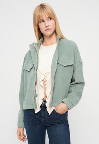 Veste en velours côtelé vert clair avec deux poches poitrine, fermeture éclair et poignets côtelés, associée à un haut crème à volants et à un jean bleu foncé.