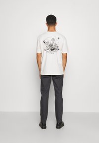 Mężczyzna odwrócony tyłem, ubrany w białą koszulkę z grafiką i czarnym napisem "Z JAPONII Z MIŁOŚCIĄ" oraz ciemne spodnie, na tle jednolitego białego tła.