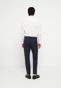 Vedere din spate a unei persoane purtând o cămașă albă cu nasturi și pantaloni eleganți bleumarin, cu pantofi negri și o textură fină. Design simplu, fără modele.