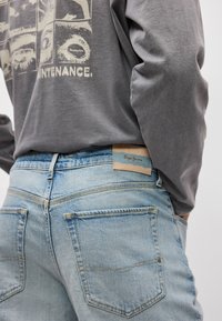 Γκρι μακρυμάνικο πουκάμισο με γραφικό σχέδιο, συνδυασμένο με ανοιχτό μπλε ξεβαμμένο τζιν που έχει πίσω τσέπες και καφέ ετικέτα με επωνυμία.