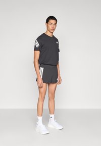 T-shirt de sport gris à manches courtes avec des bandes blanches sur les épaules, assorti à un short gris. Des baskets blanches et des chaussettes chevilles complètent la tenue.