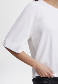 Witte gebreide trui met korte mouwen, voorzien van een geribde zoom en subtiele textuur. Geen zichtbare patronen; gladde stof met minimale ontwerpdetails.