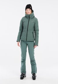 Groene geïsoleerde skijas met een hoge kraag, zwarte rits en verborgen zakken. Gecombineerd met bijpassende groene skibroek en zwarte skischoenen.