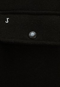 Black wool fabric with a silver "J" logo and a shiny black button featuring "BUTCHER" engraved around the edge.