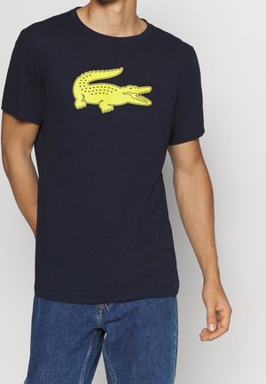 Mężczyzna ubrany w granatowy, krótki rękaw, T-shirt z dużym, jaskrawożółtym logo krokodyla, w połączeniu z niebieskimi dżinsami.