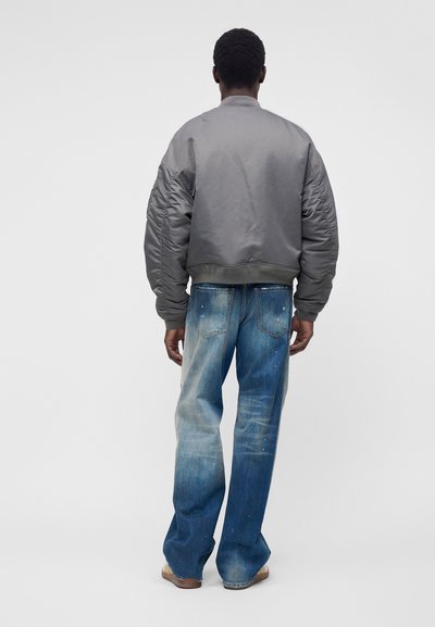 Homme de dos portant une veste bomber grise, un jean bleu délavé ample avec des éclaboussures de peinture et des chaussures marron.