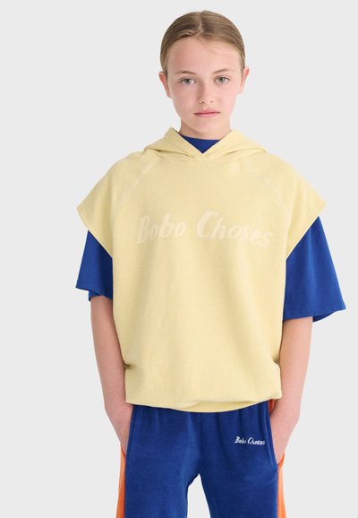 Jeune personne les mains dans les poches, portant un sweat à capuche sans manches jaune sur un chemisier bleu et un pantalon bleu avec des rayures orange.