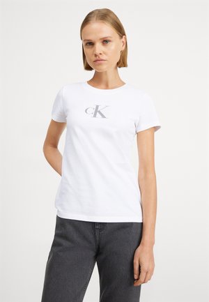 T-shirt en coton blanc avec un col rond, des manches courtes et un logo "CK" gris au centre. Le modèle porte un jean gris.