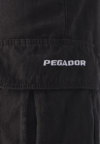 Pantaloni neri in tessuto con cuciture a vista, dotati di un logo ricamato "PEGADOR" in bianco nella zona della tasca anteriore.