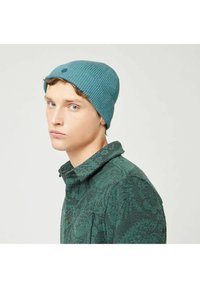Teal geribbelde trui met een kleine geborduurde stip, gedragen met een gestreepte groene button-up shirt, zijwaarts tentoongesteld.