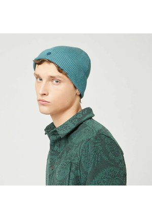 Teal ribbed knit beanie with a small embroidered dot, worn with a patterned green button-up shirt, showcased from the side.