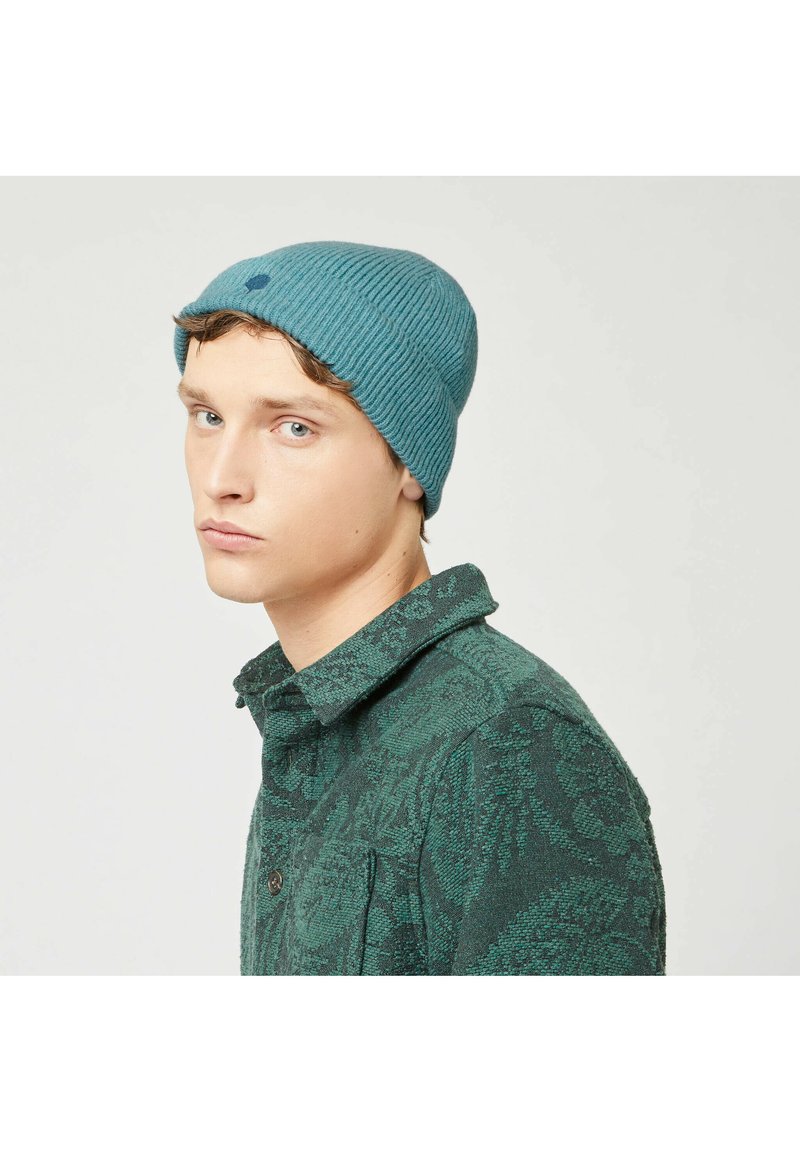 Berretto a coste color teal con un piccolo punto ricamato, indossato con una camicia verde a fantasia, mostrato di lato.